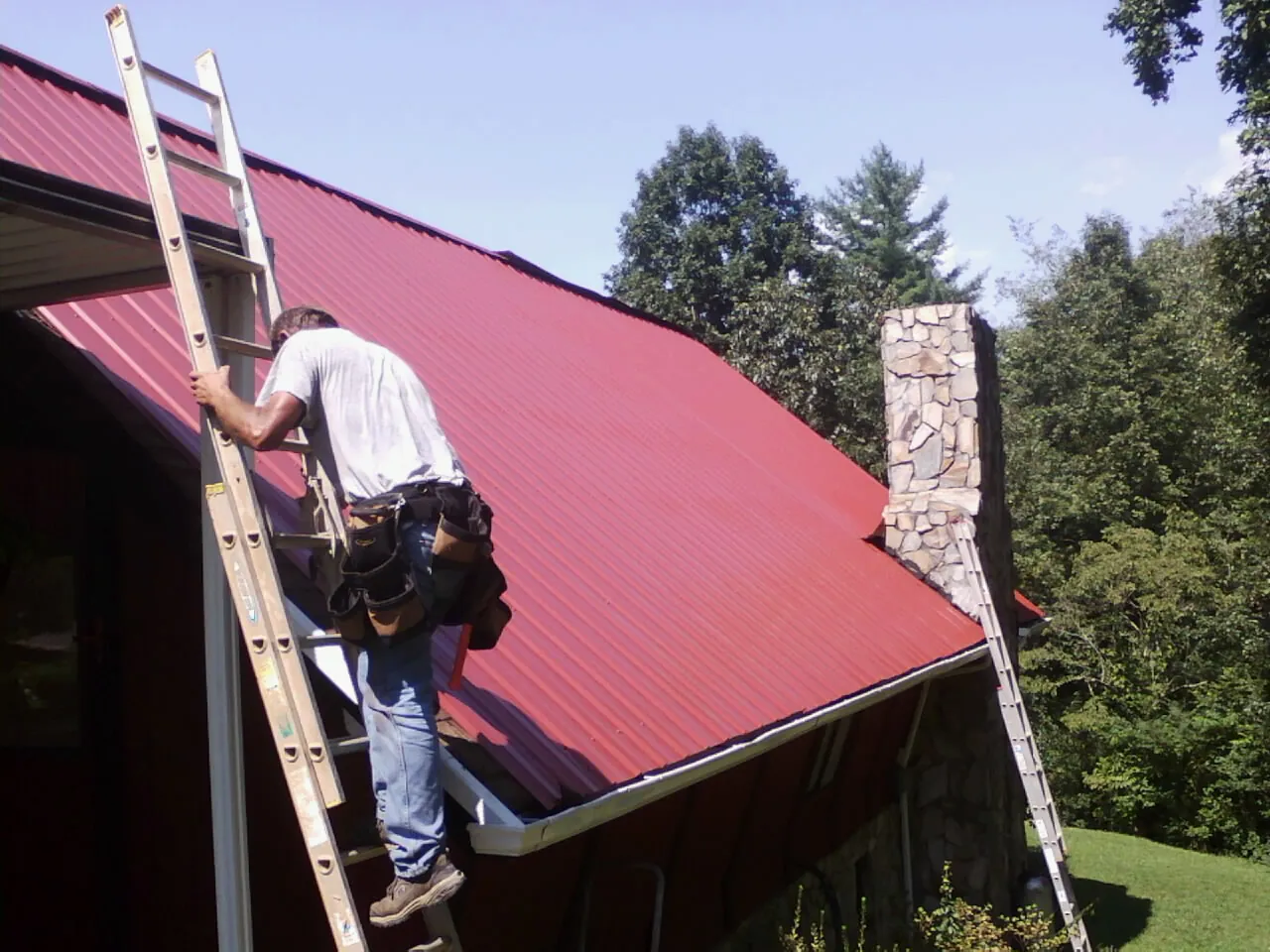 Roofer on a ladder beside a red metal roof and stone chimney — Soffit & Fascia Repair by Roofing Brothers in Laurys Station